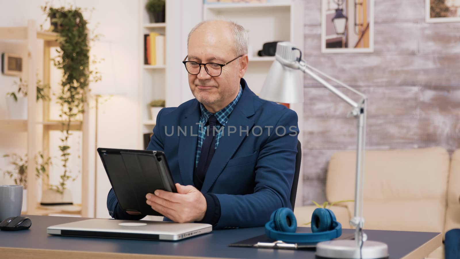 Elederly man using tablet while sitting down at the office in living room. Elderly woman walking in the background