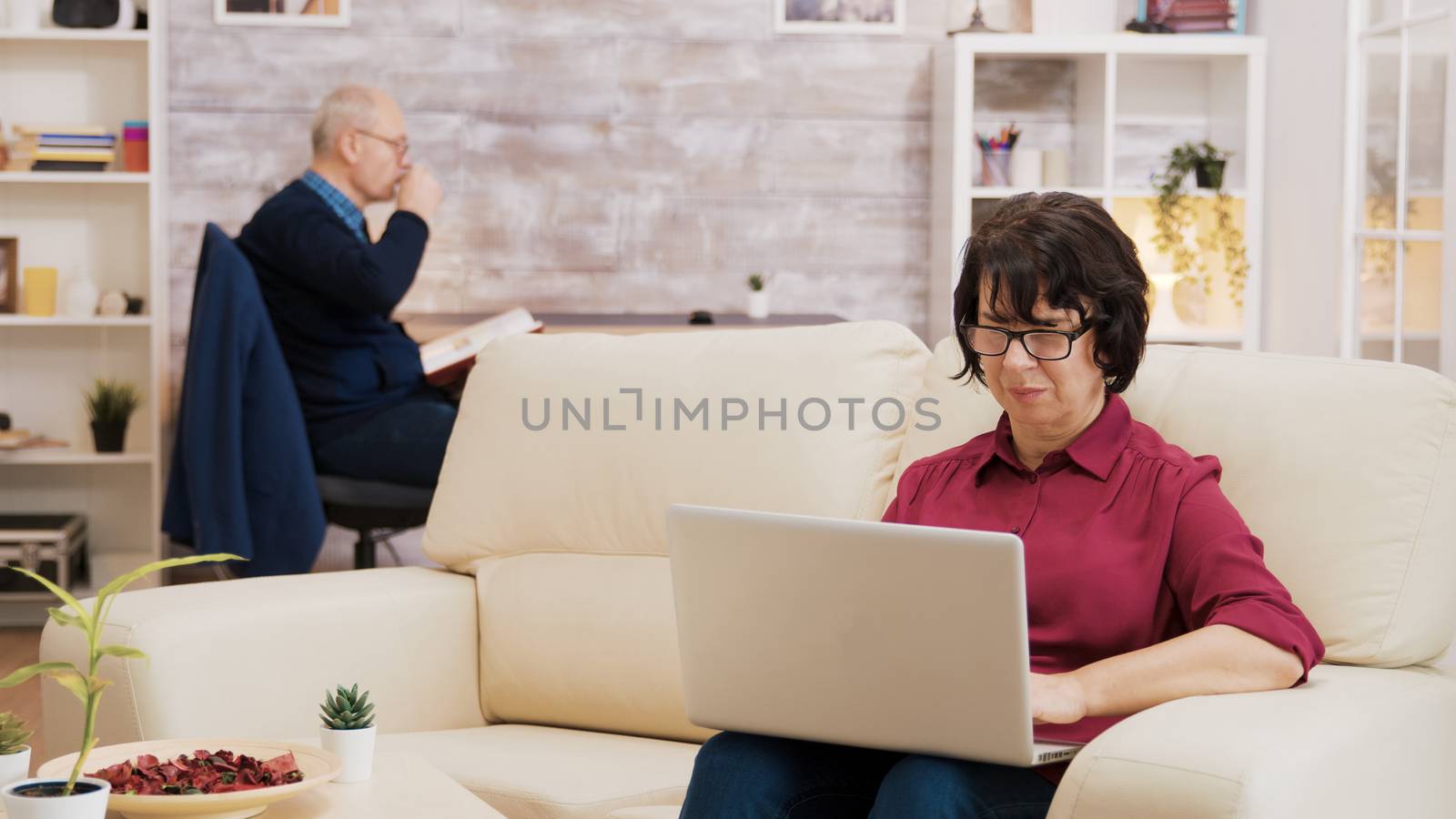 Senior woman with glasses working on laptop sitting on sofa. Elderly age man reading a book in the background.