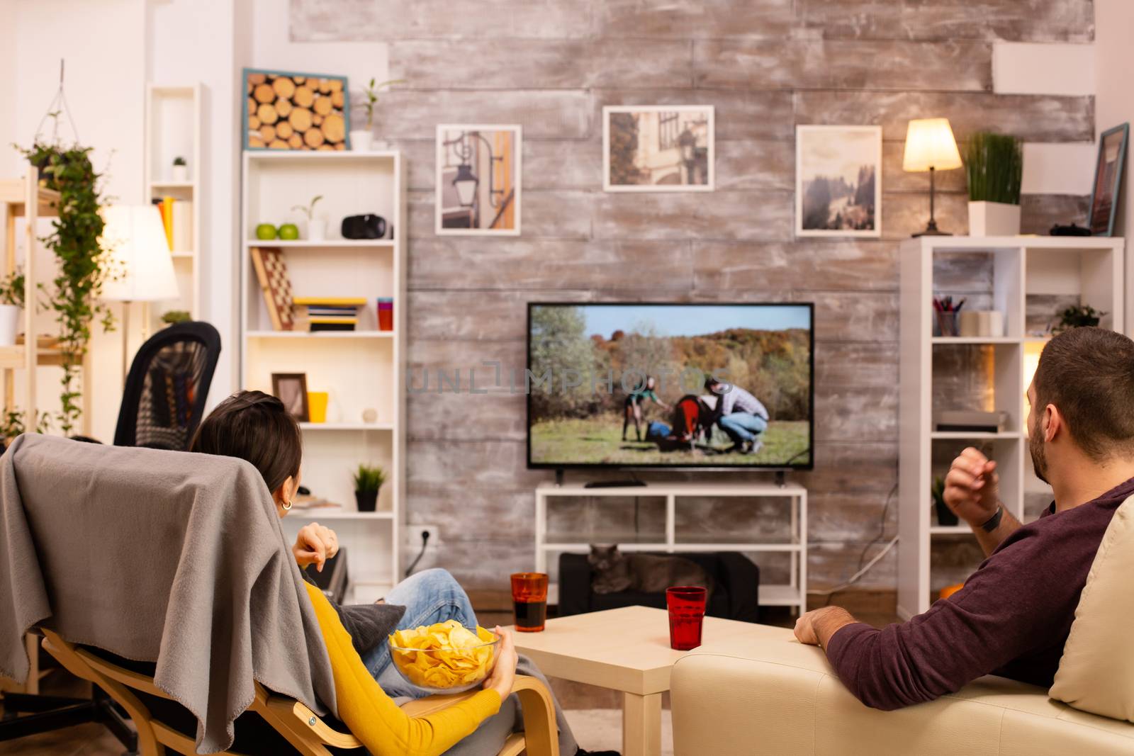 Back view of couple in living room watching a movie on the TV while eating takeaway food