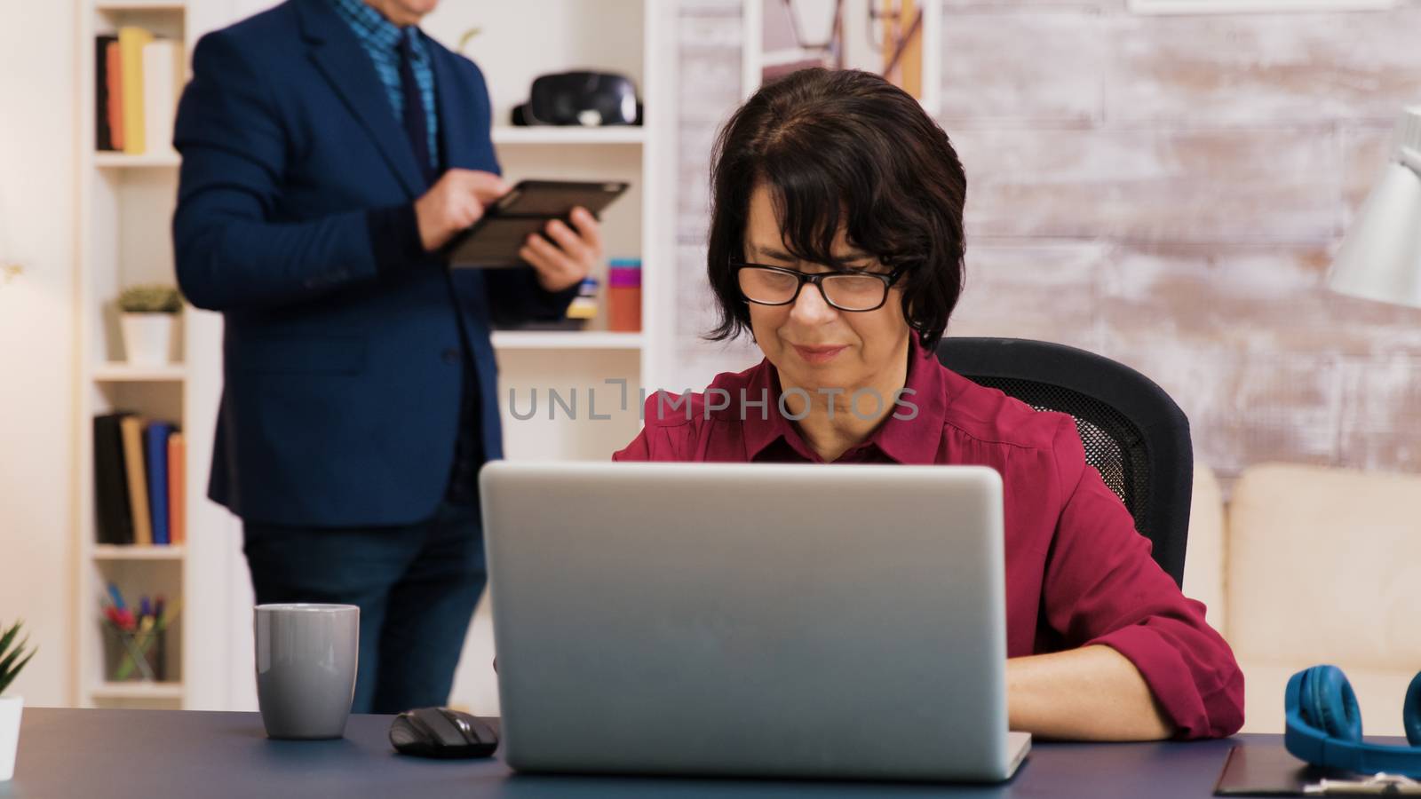 Retired woman working on laptop in living room while her husband is using tablet in the background.