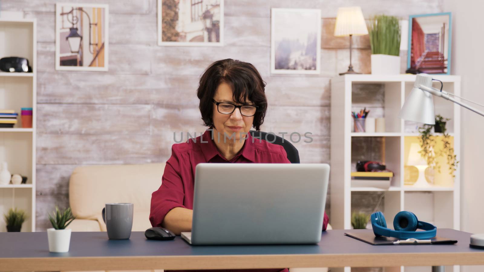 Elderly age woman taking a sip of coffee while browsing on laptop. Old man relaxing on sofa in the background.