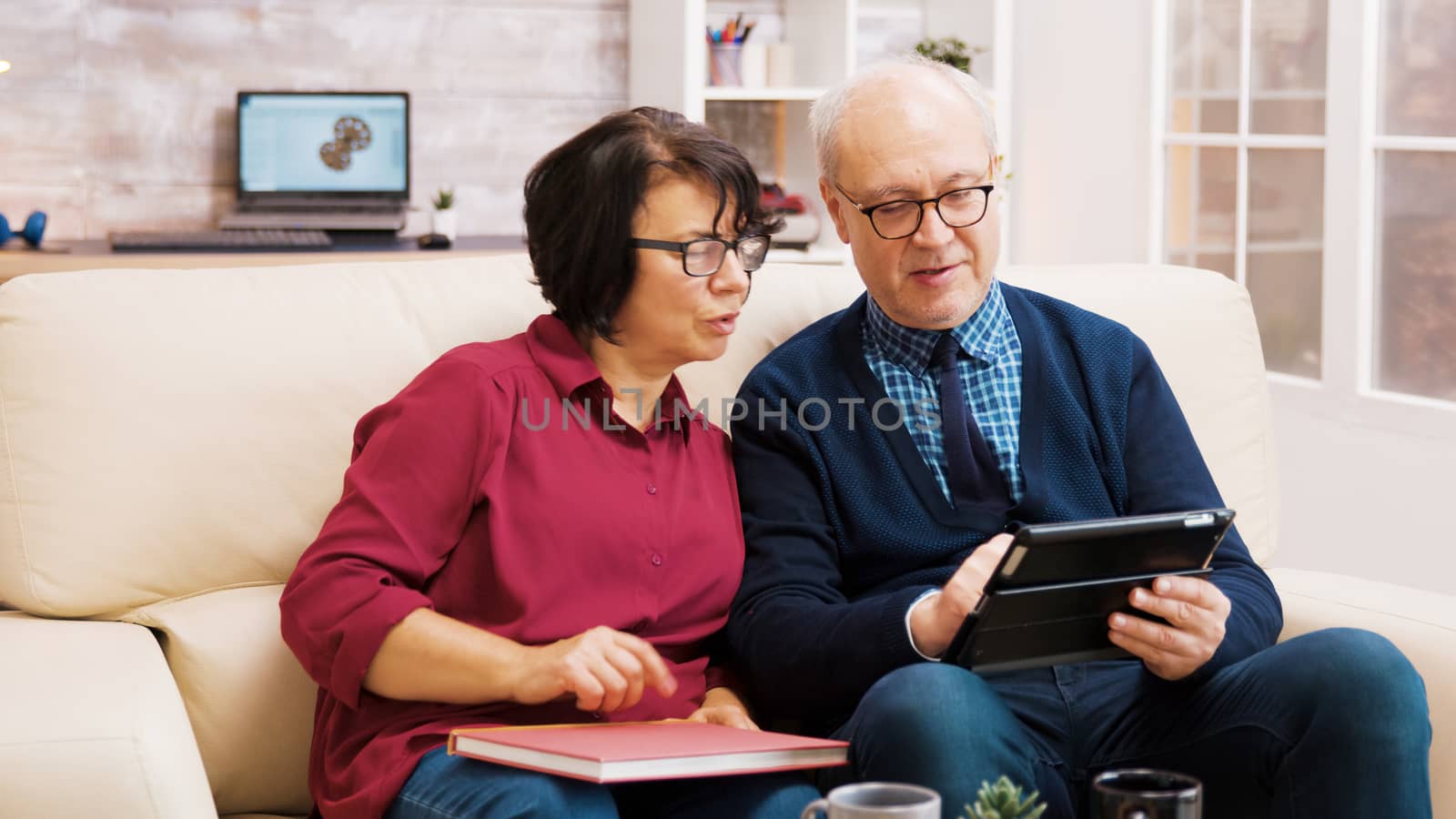 Elderly age couple using tablet while sitting on sofa in living room. Elderly age woman enjoying a cup of coffee.