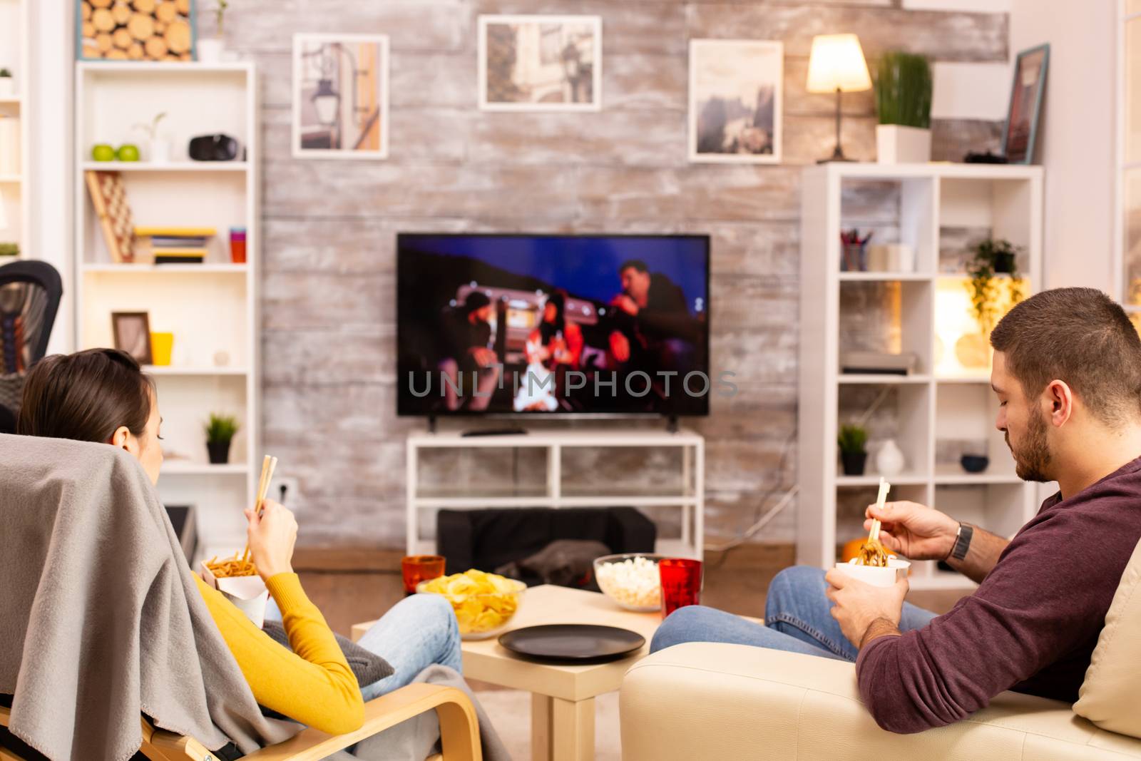 Back view of couple in living room watching a movie on the TV while eating takeaway food