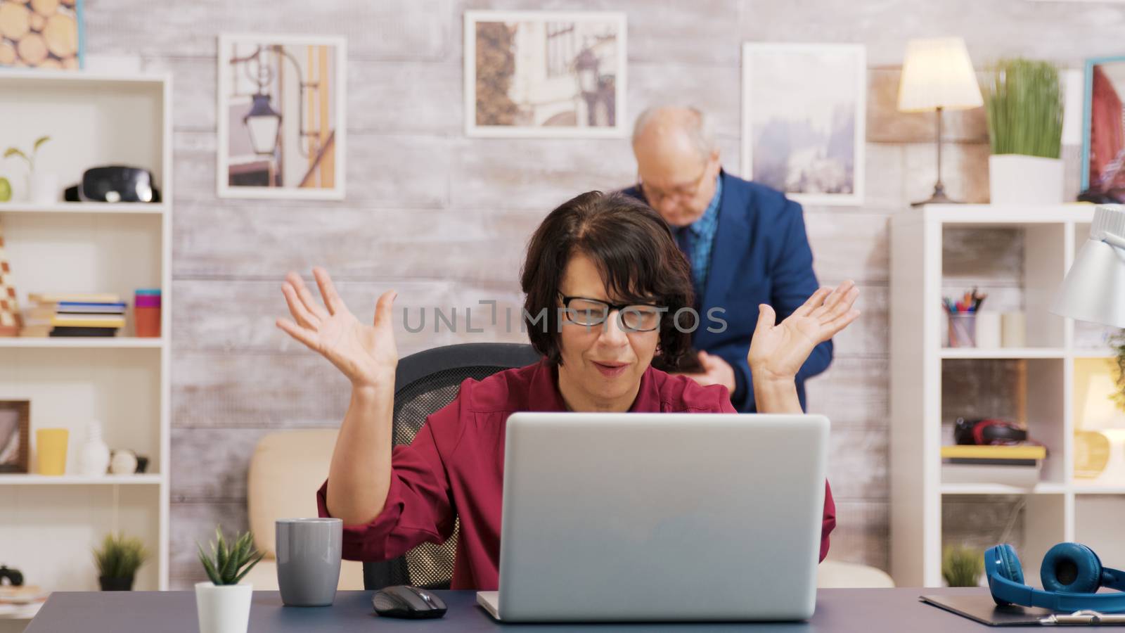 Amazed old woman after reading a news on laptop in living room. Old man using tablet sitting on sofa in the background.