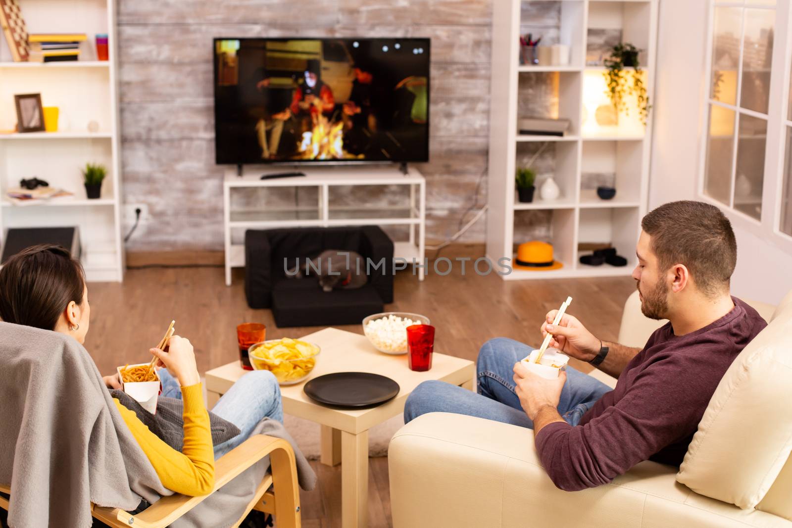 Back view of couple in living room watching a movie on the TV while eating takeaway food