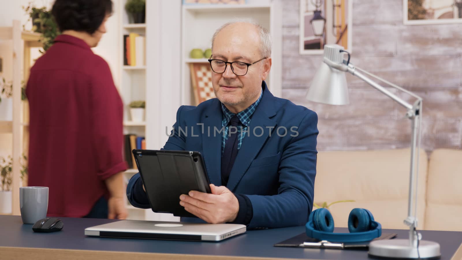 Elederly man using tablet while sitting down at the office in living room. Elderly woman walking in the background