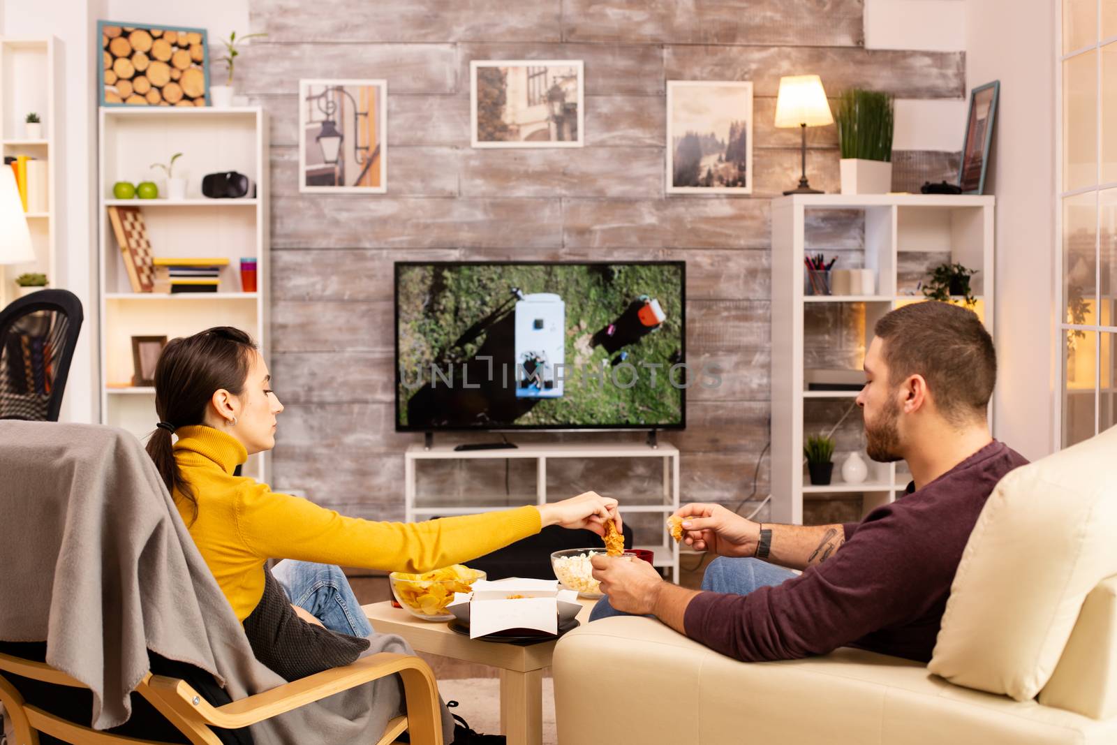 Back view of couple in living room watching a movie on the TV while eating takeaway food