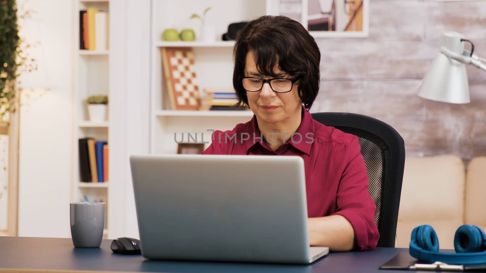 Retired woman working on laptop in living room while her husband is using tablet in the background.