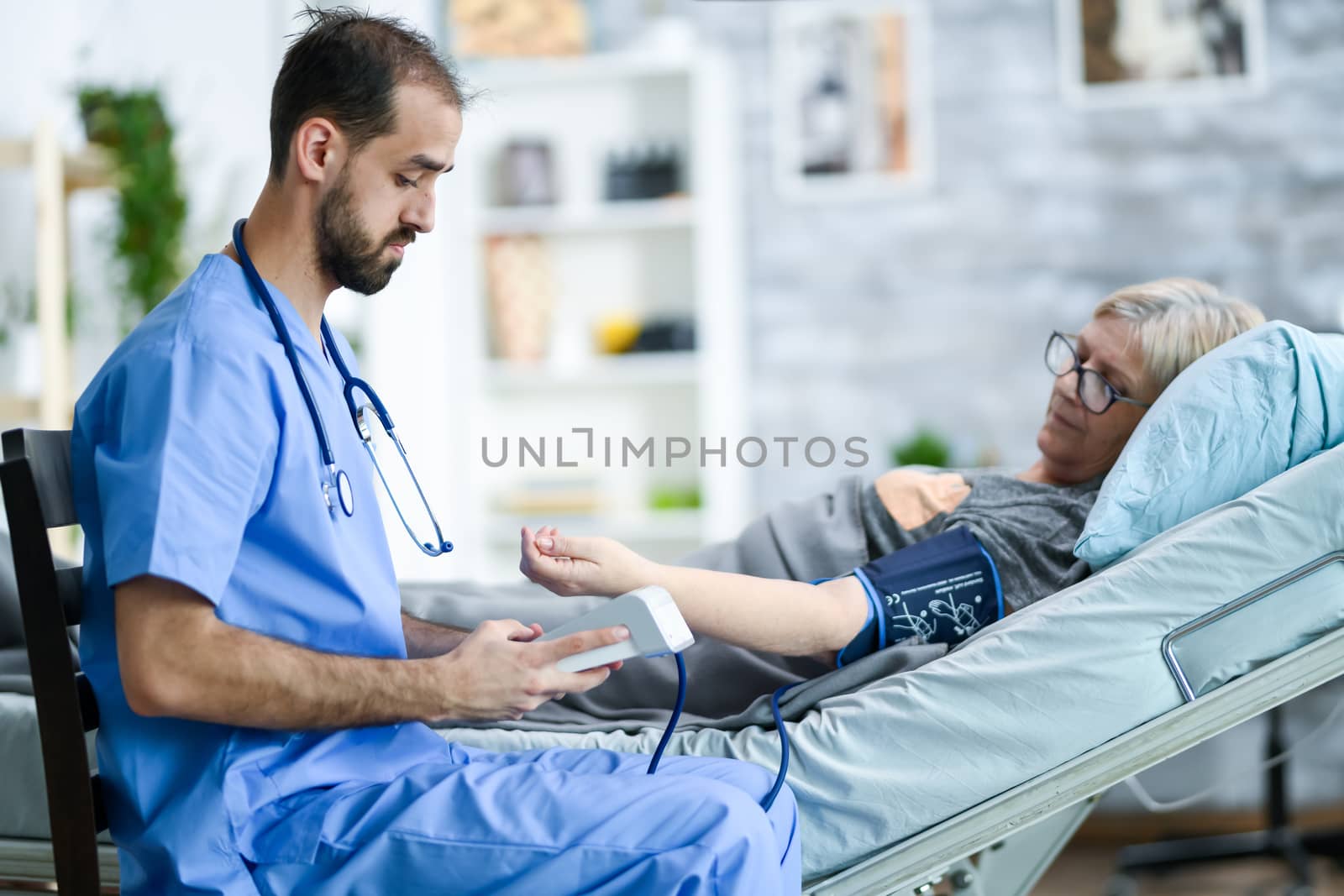 Young doctor with stethoscope in nursing home doing examination of blood pressure of a old woman using digital device.