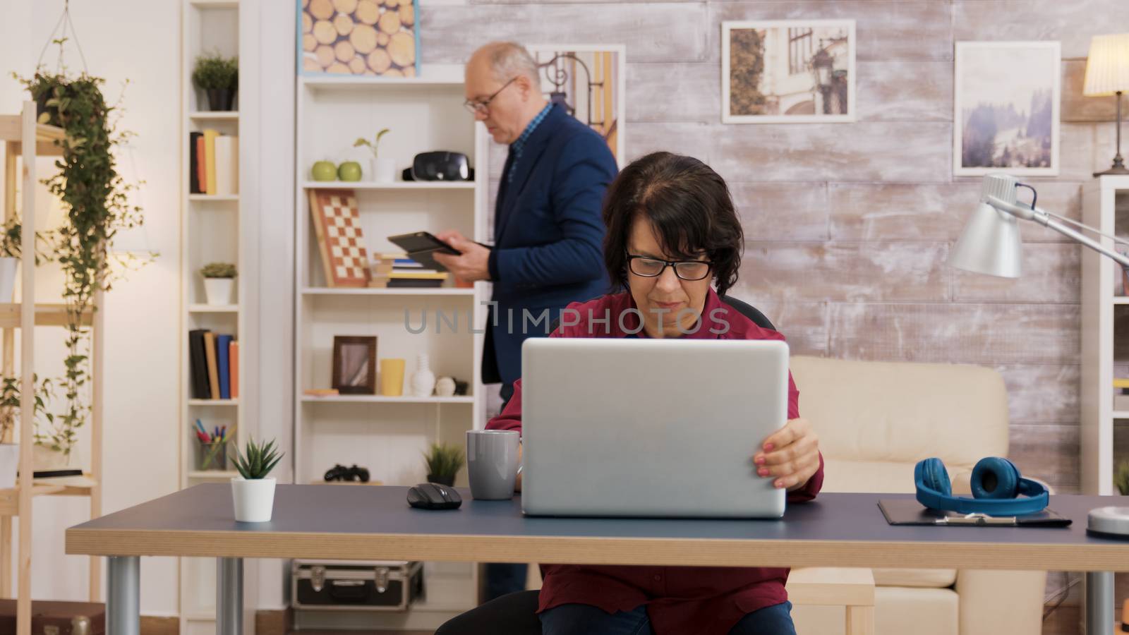 Elderly woman opening laptop and putting his glasses on her face. Old man with glasses using tablet in the brackground.