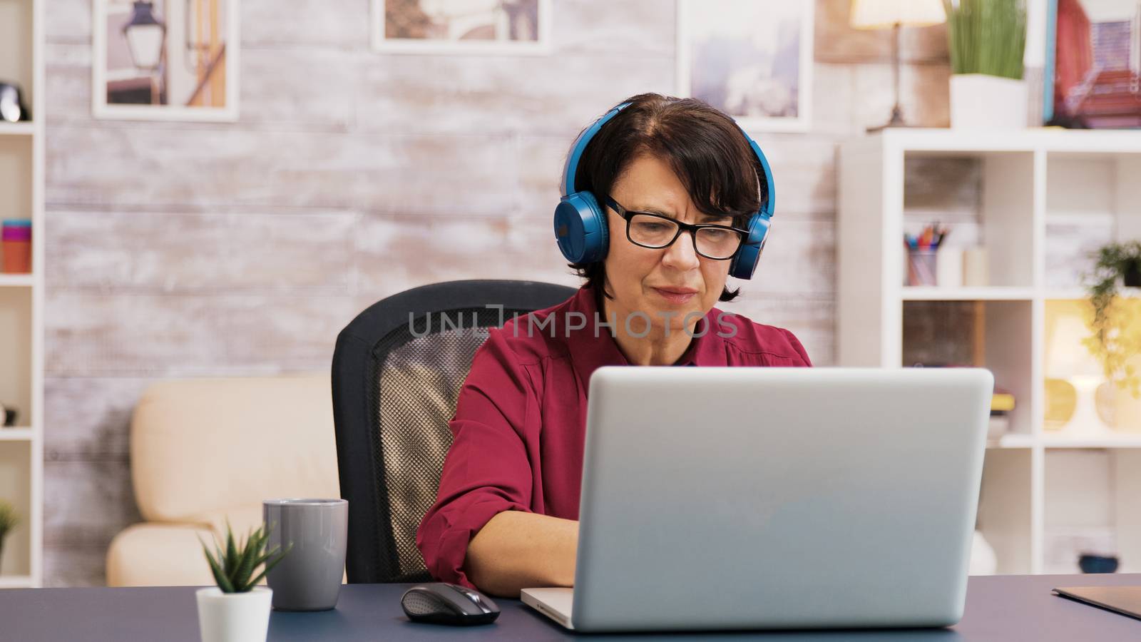 Old woman enjoying a cup of coffee while working on laptop with headphones on her head. Elderly man using tablet in the background.