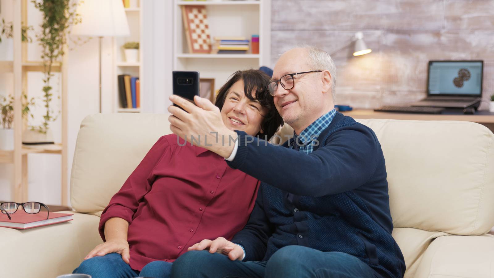 Happy senior couple sitting on sofa taking a selfie by DCStudio