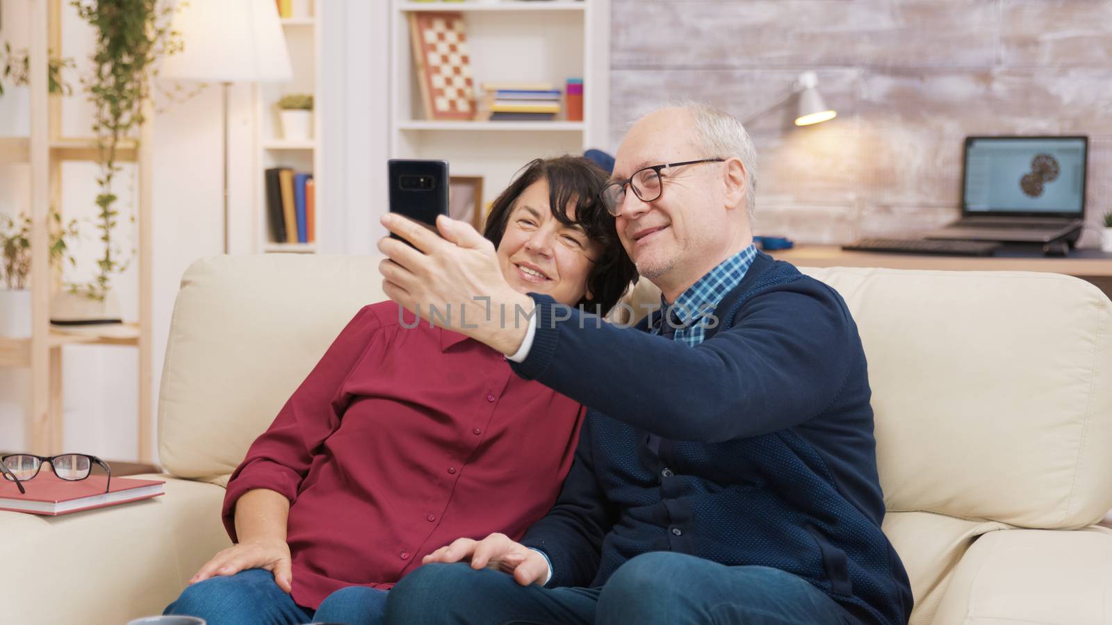 Happy senior couple sitting on sofa taking a selfie in the living room