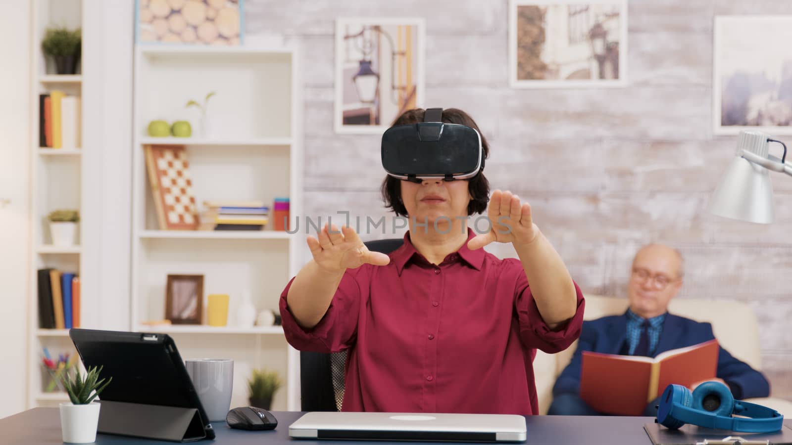 Retired senior woman amazed while using virtual reality goggles in living room. Elderly man sitting on sofa while reading a book.