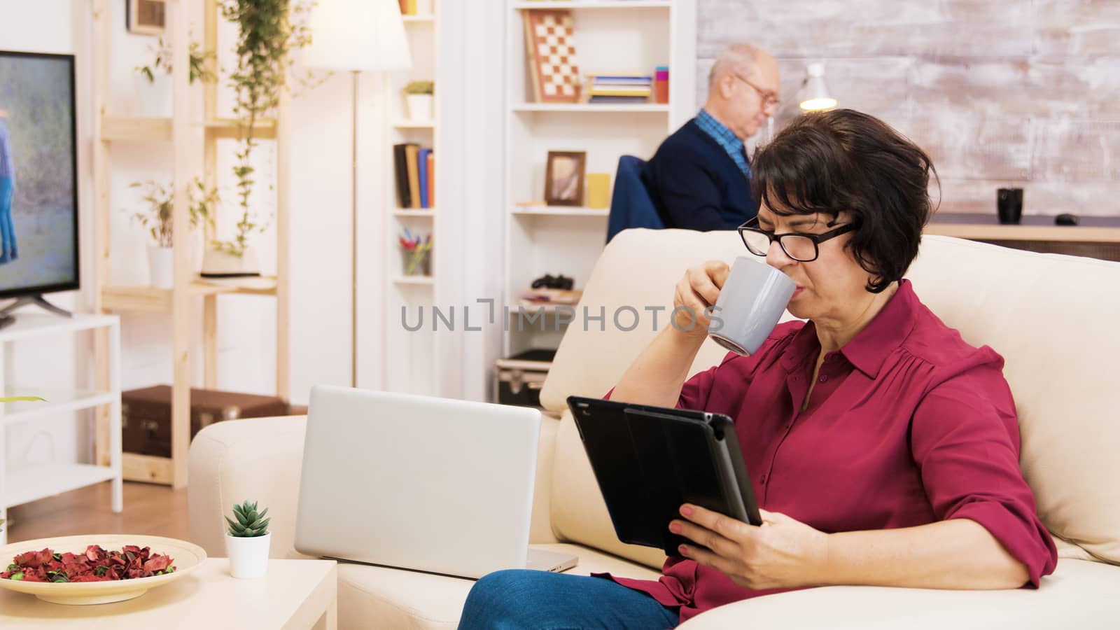 Zoom in shot of elderly age woman using tablet sitting on coich. Senior man reading a book in the background.