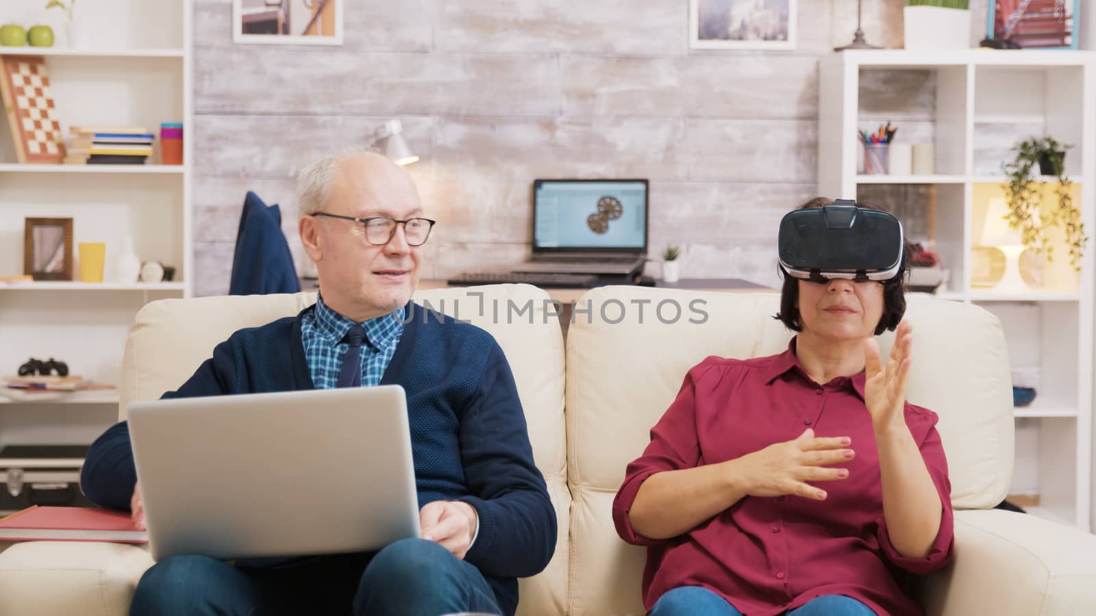 Elderly age woman sitting on sofa wearing virtual reality goggles. Old man sitting on sofa using laptop next to his wife