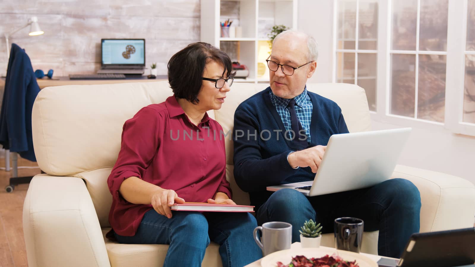 Elderly age couple using laptop while sitting on sofa in living room. Elderly woman taking a sip of coffee.