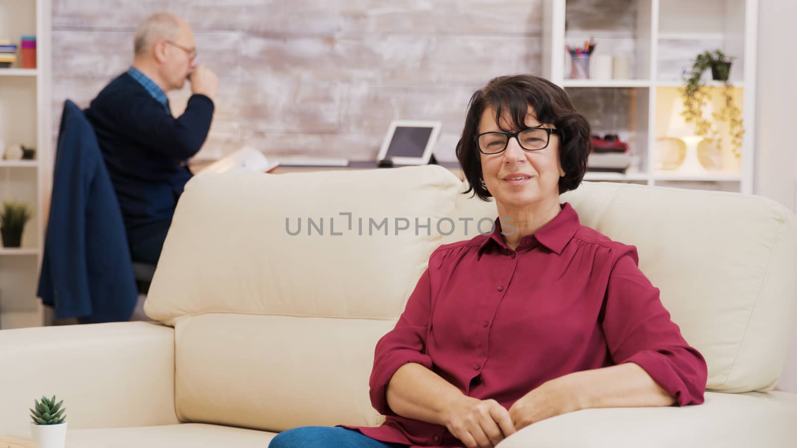 Portrait of elderly age woman sitting on sofa putting glasses on by DCStudio