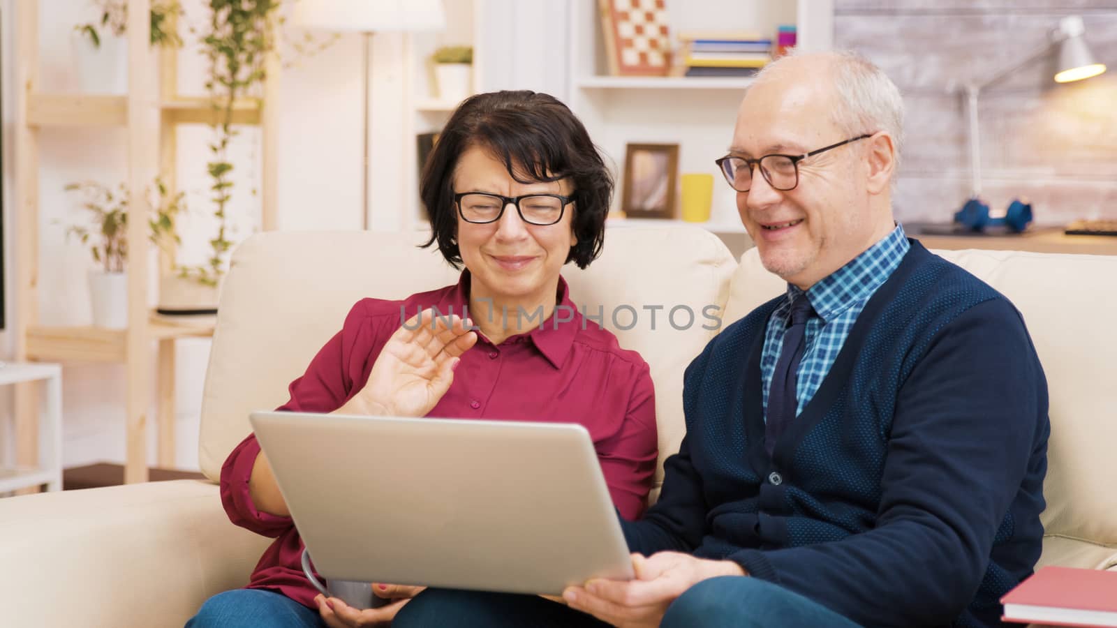 Elderly age couple sitting on sofa holding laptop during a video call. Couple waving at laptop.