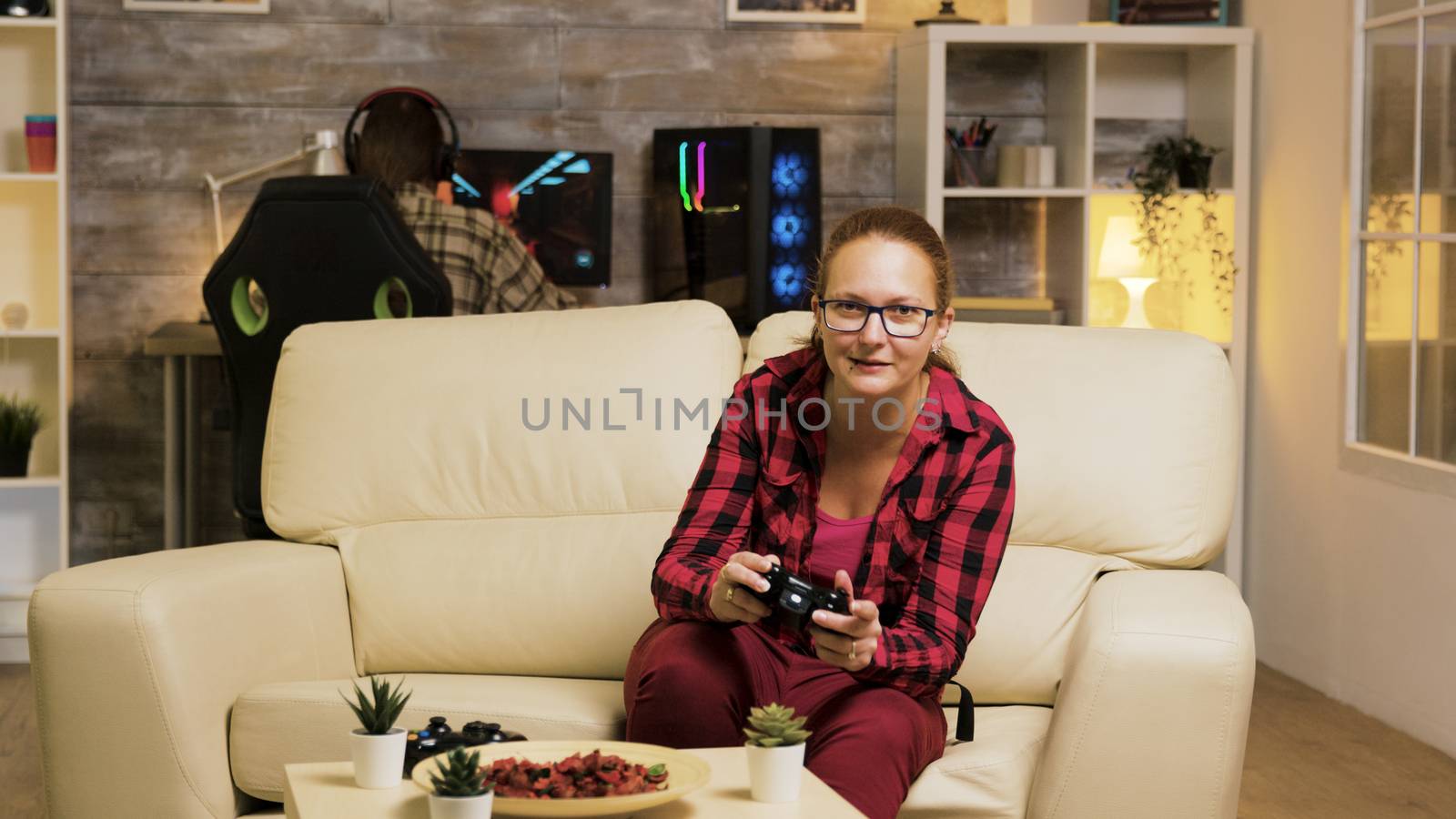Woman sitting on couch in living room playing video games using wireless controllers. Boyfriend playing games on computer in the background.