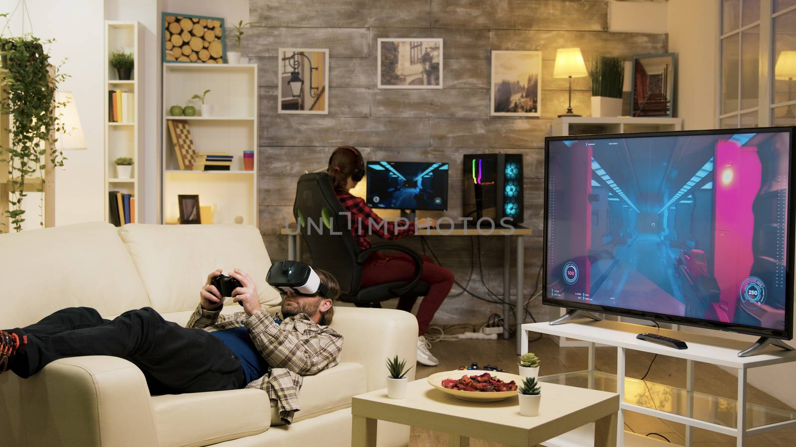 Man lying on sofa playing video games using vr headset with his girlfriend in the background playing on computer.