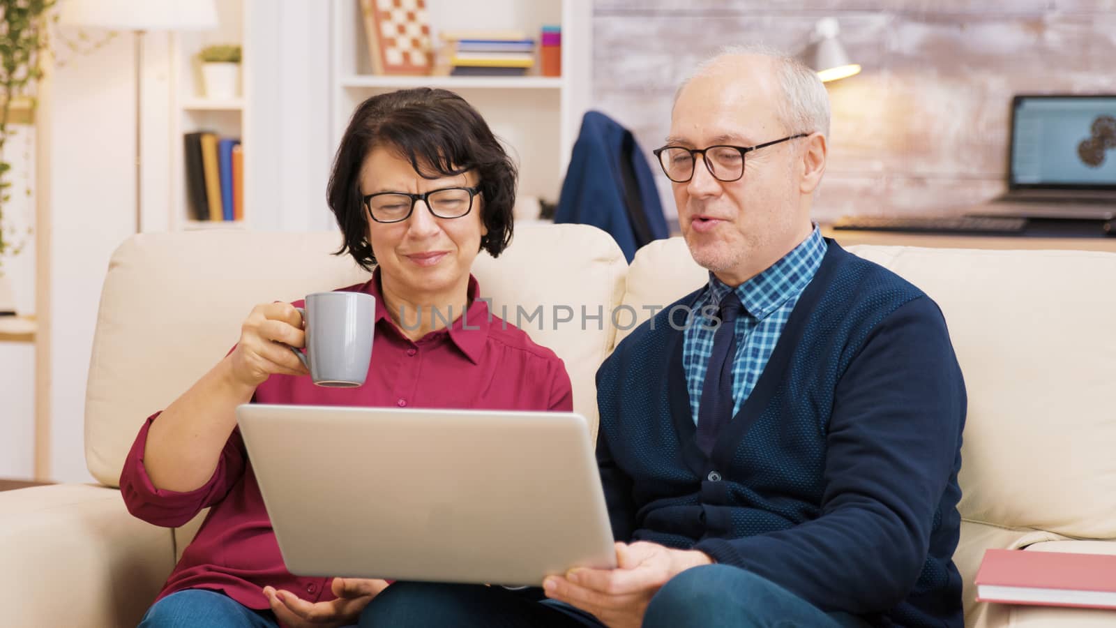 Elderly age couple sitting on sofa holding laptop during a video call. Couple waving at laptop.