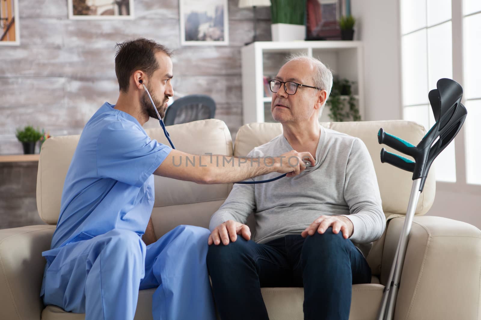 Young male nurse with stethoscope listening the heart of senior man in nursing home.