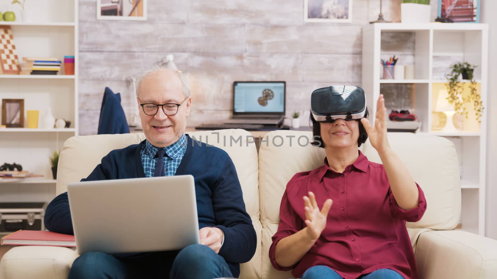 Elderly age woman sitting on sofa wearing virtual reality goggles. Old man sitting on sofa using laptop next to his wife