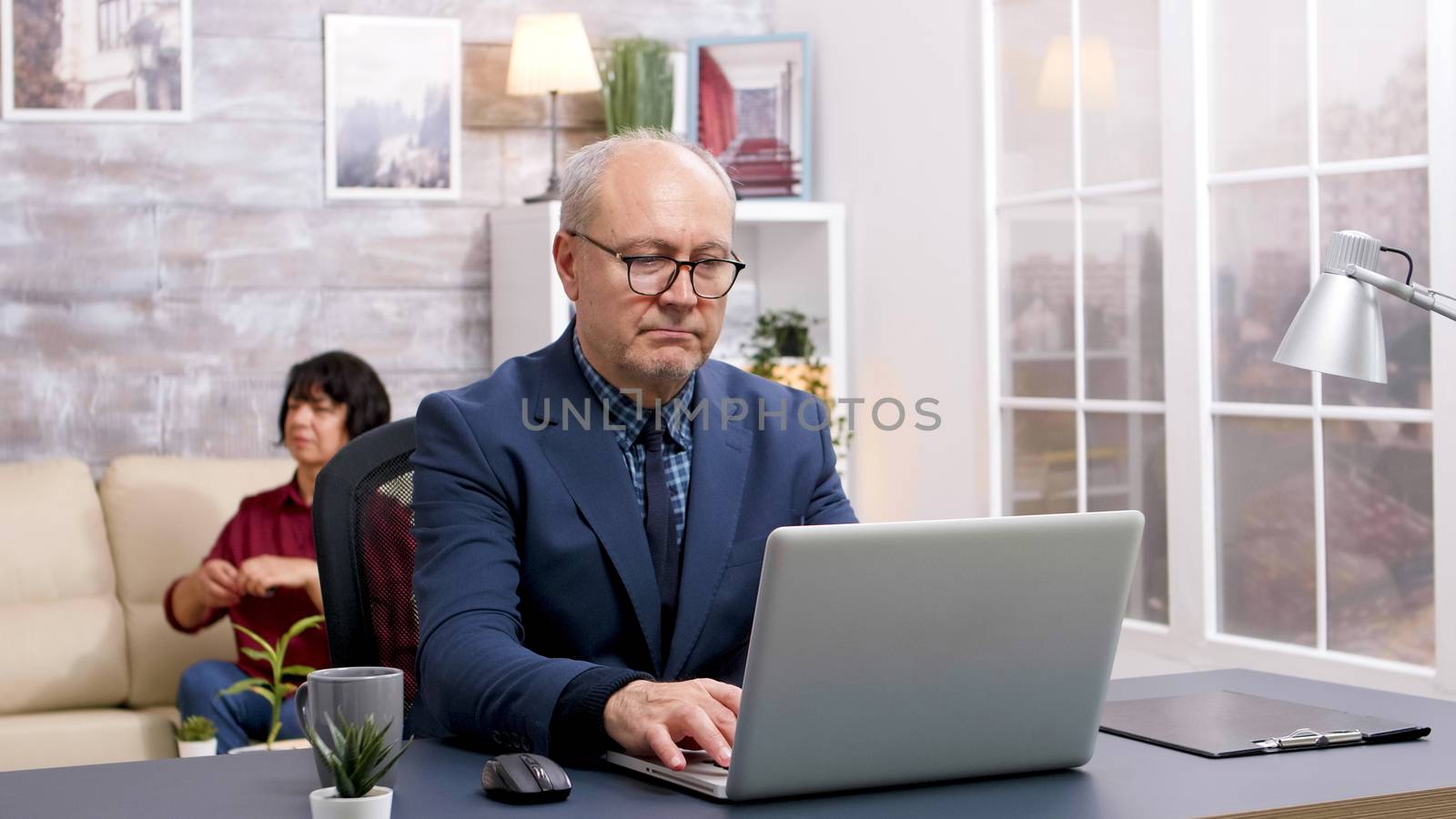 Old man working on laptop and taking a sip of coffee in living room with wife sitting on sofa in the background.