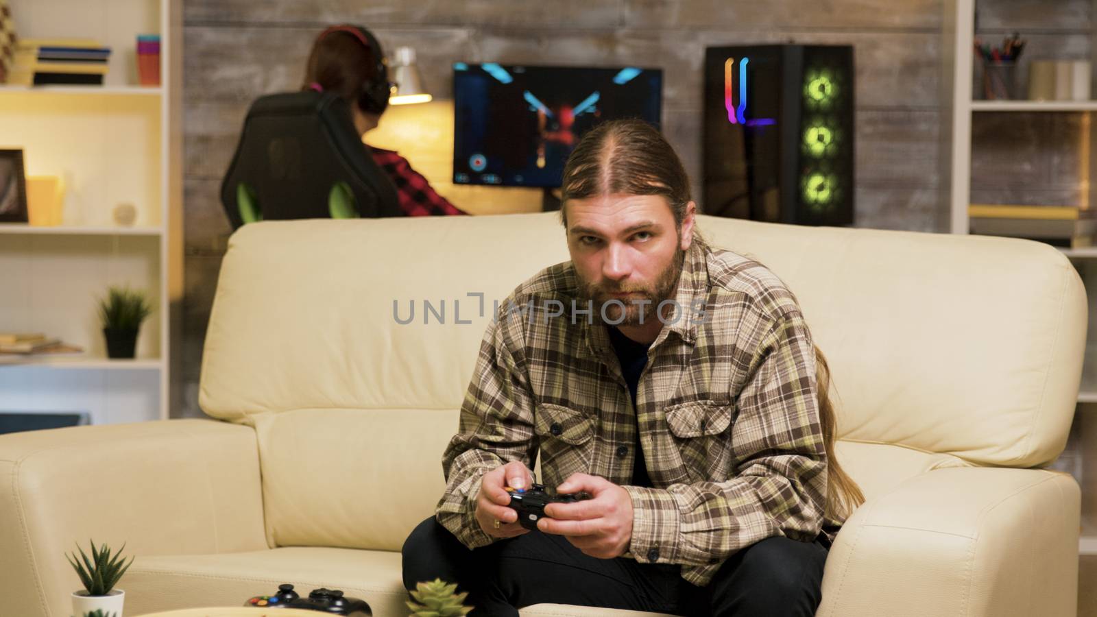Focused bearded man sitting on couch playing video games using wireless controller. Girlfriend playing on computer in the background.