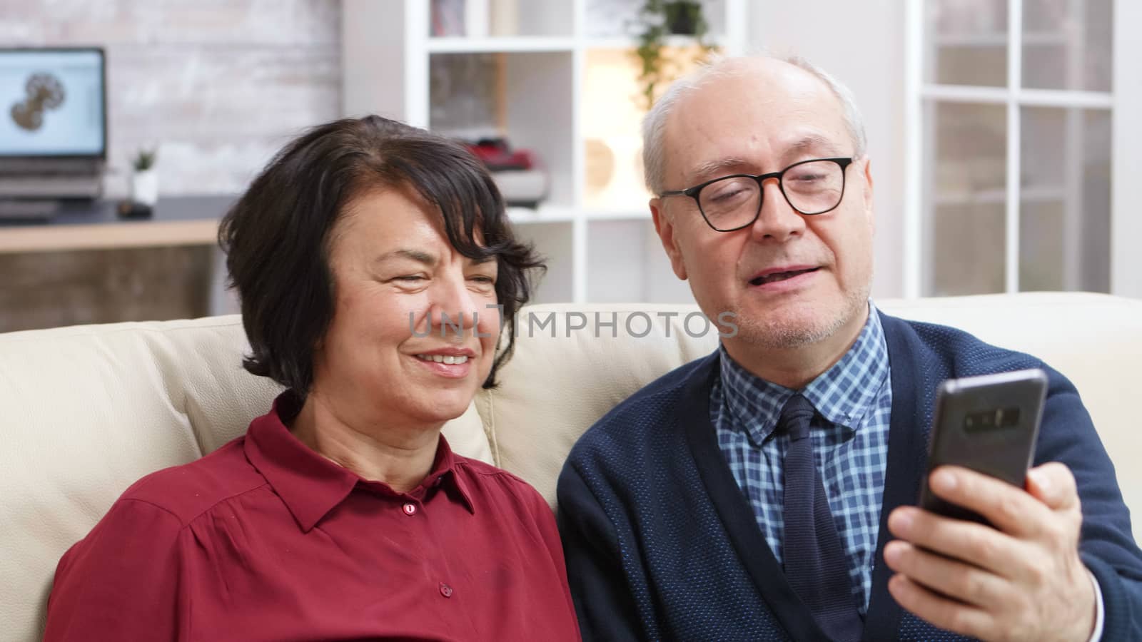Cheerful old couple sitting on sofa taking a selfie in living room.