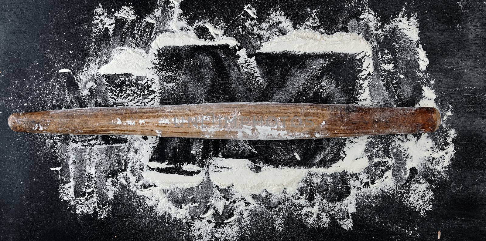 very old wooden rolling pin on a black background, top view, scattered white wheat flour