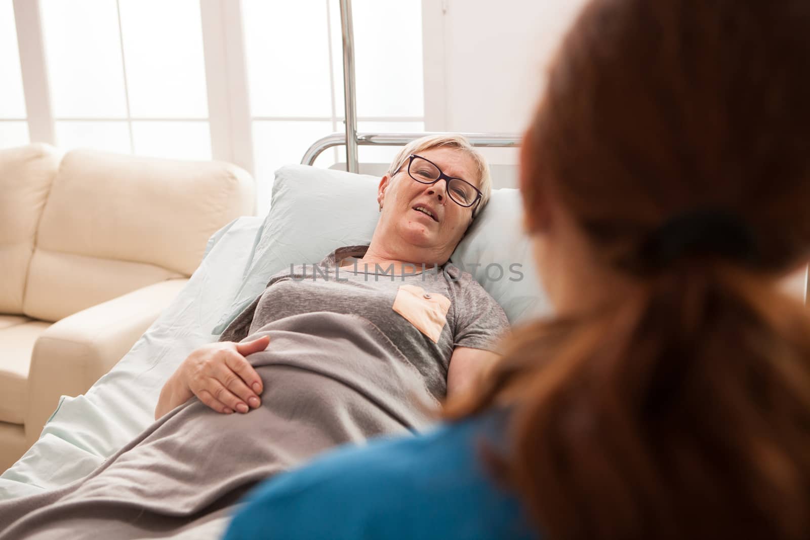 Back view of female doctor talking with senior woman laying in bed in a nursing home.