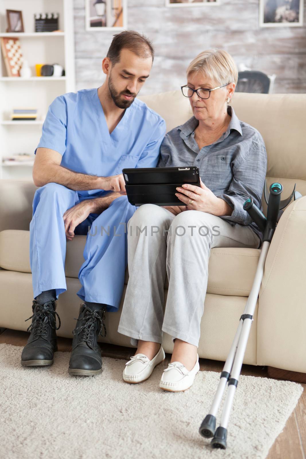 Senior woman with crutches in nursing home using tablet with help from young assistant.