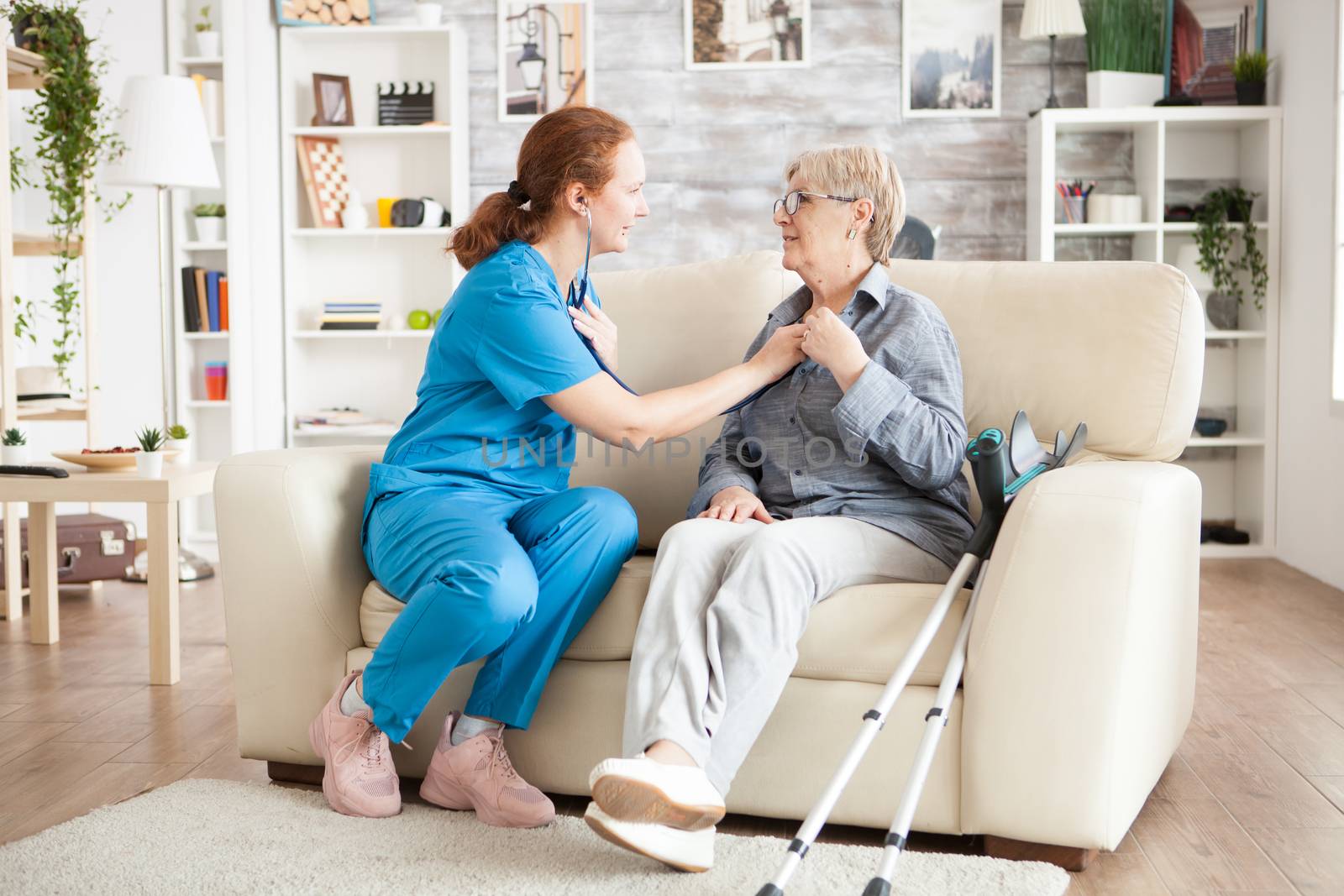 Female nurse sitting on couch with old woman in nursing home using stethoscope to listen her heartbeat.