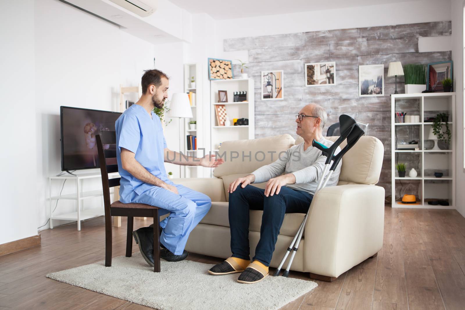 Male nurse on a chair in a nursing home talking with elderly age man while he is sitting on couch.