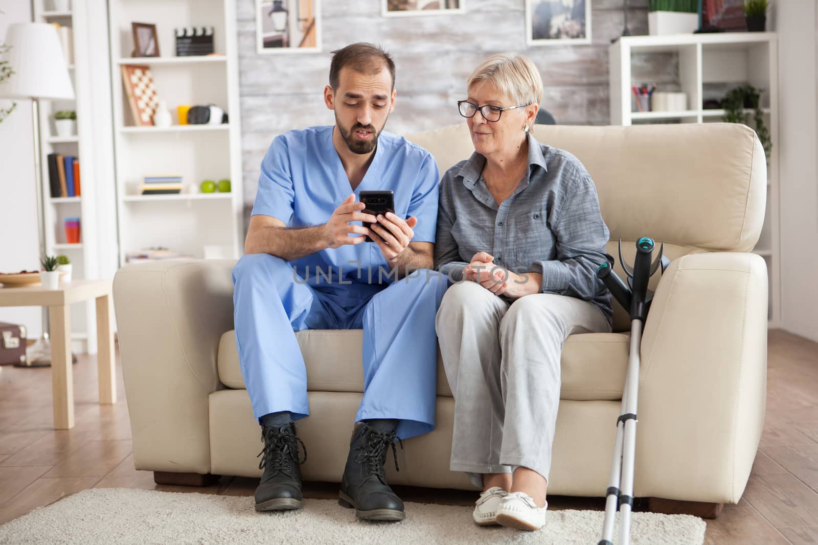 Young male doctor in nursing home helping senior woman to use mobile phone.