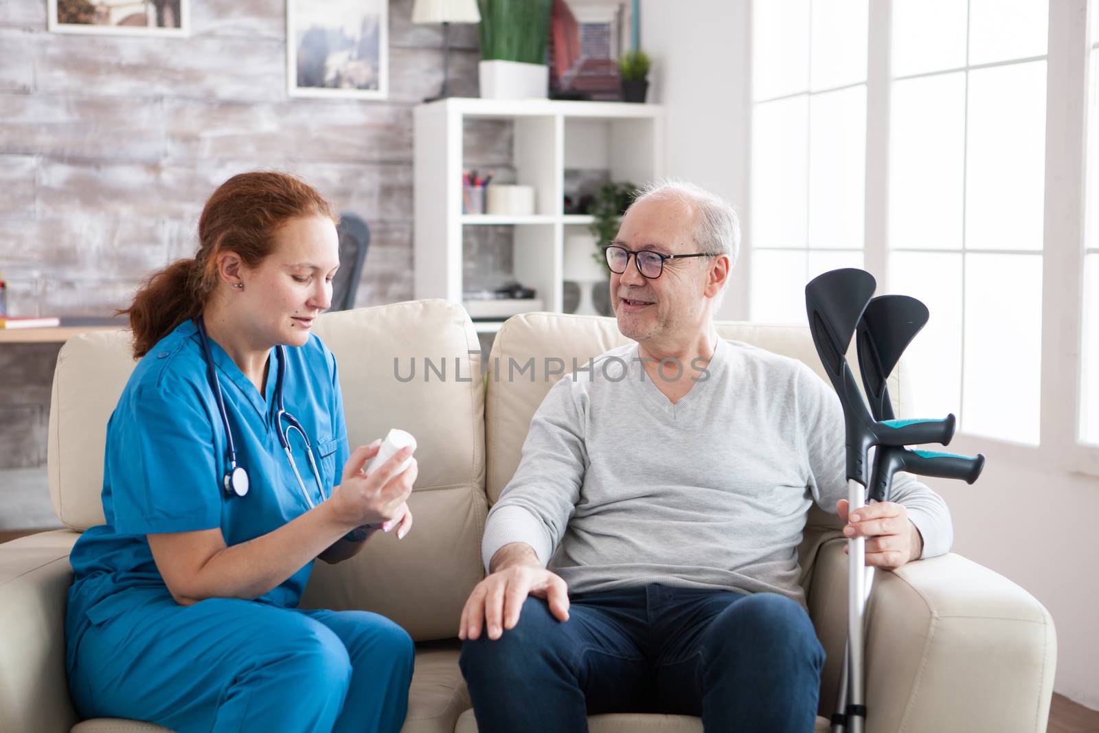 Female care taker telling cheerful senior man with crutches in nursing home how to take his pills.