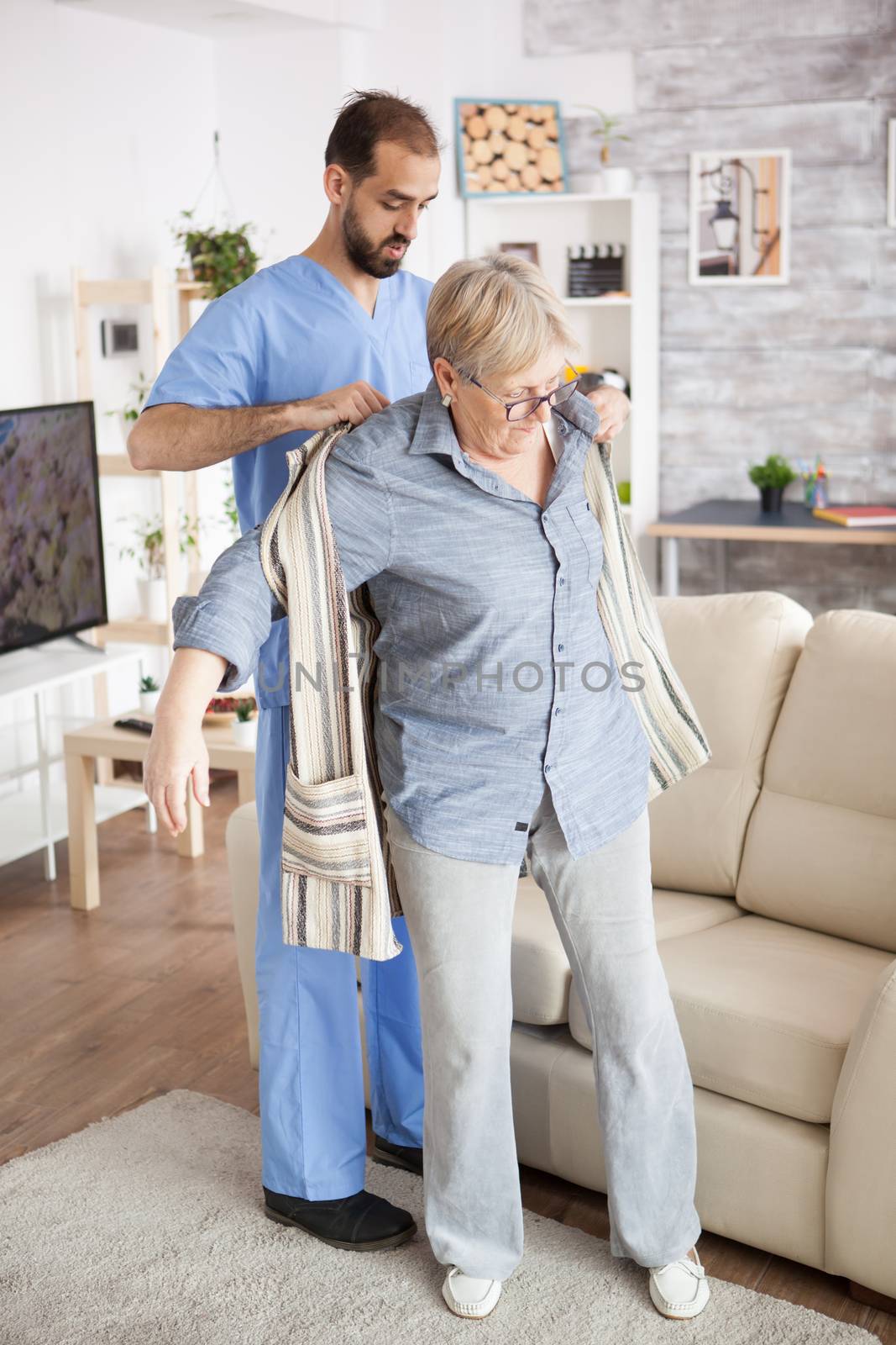 Young male caregiver in nursing home helping senior woman getting dressed.