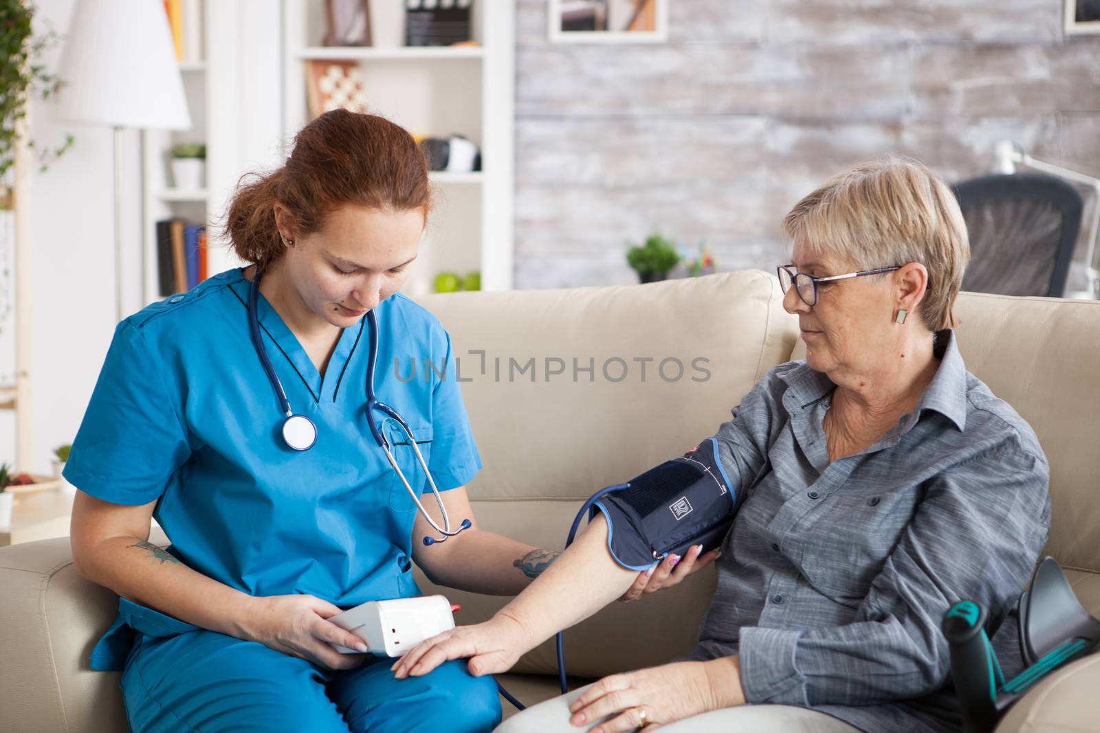 Nurse sitting on couch with old woman in nursing home checking her blood pressure with digital device.