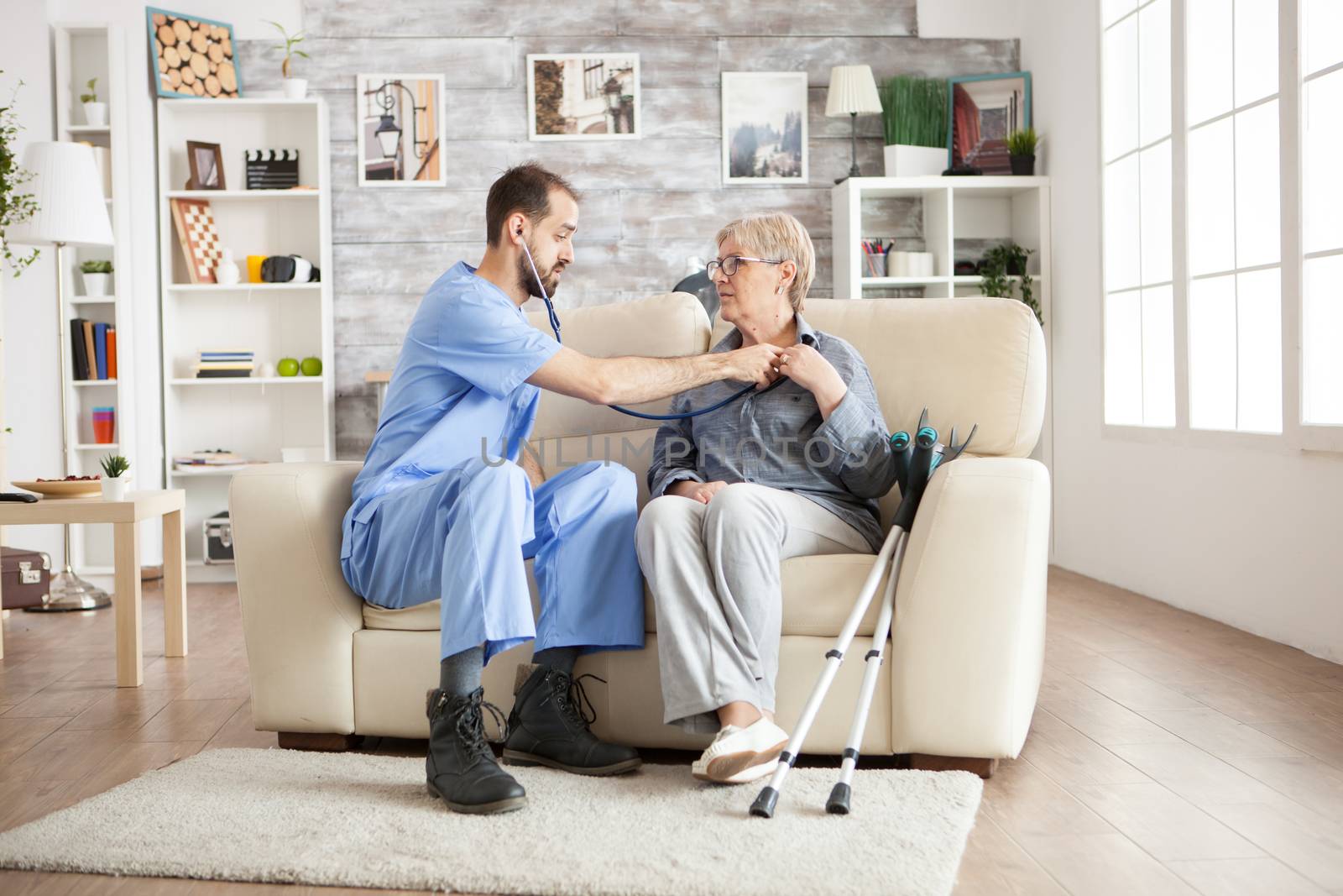 Doctor with stethoscope in a nursing home checking old woman heart beat sitting on couch. Old woman with crutches.
