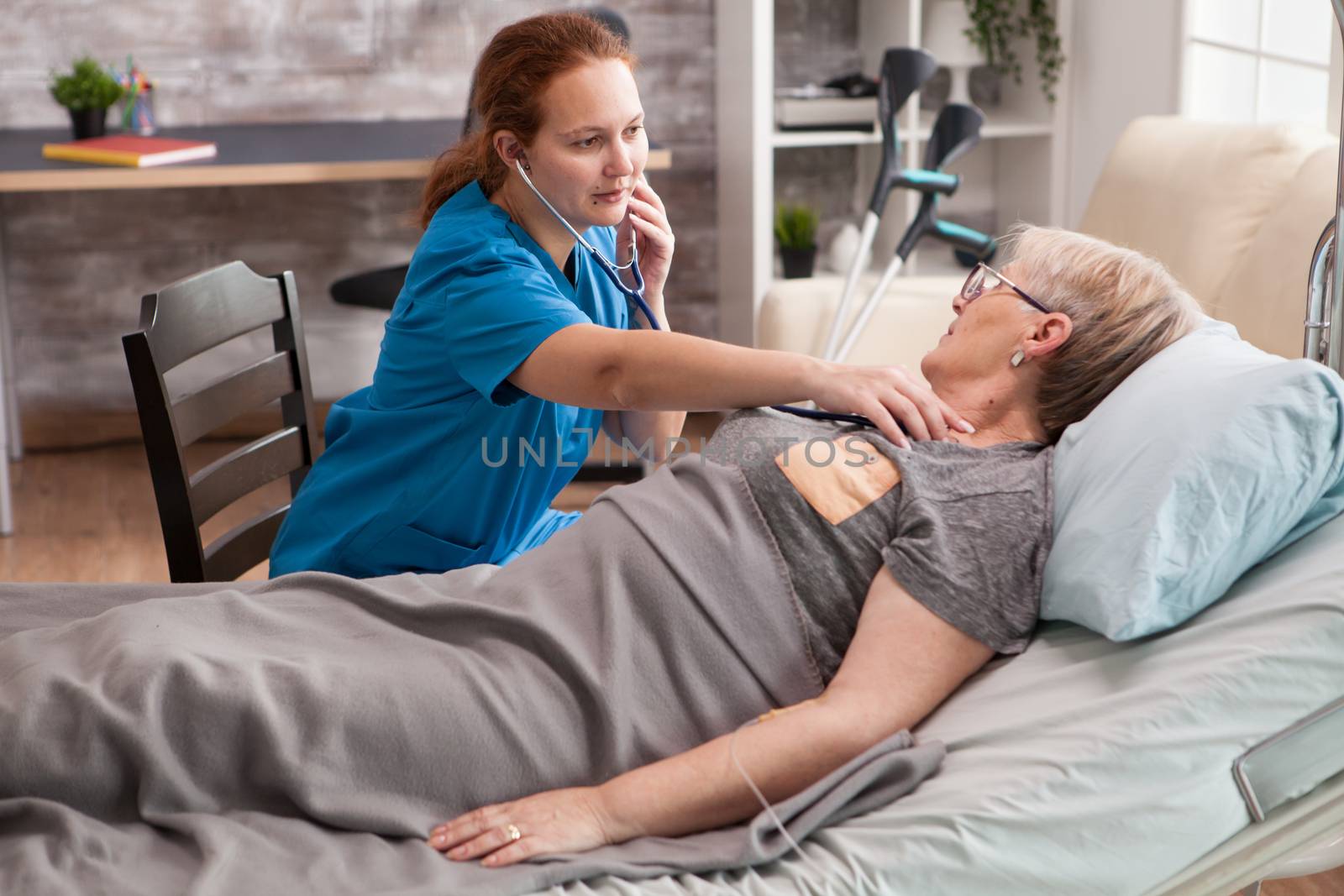 Female nurse in nursing home doing hear beat mesasurement of old woman sitting on bed.
