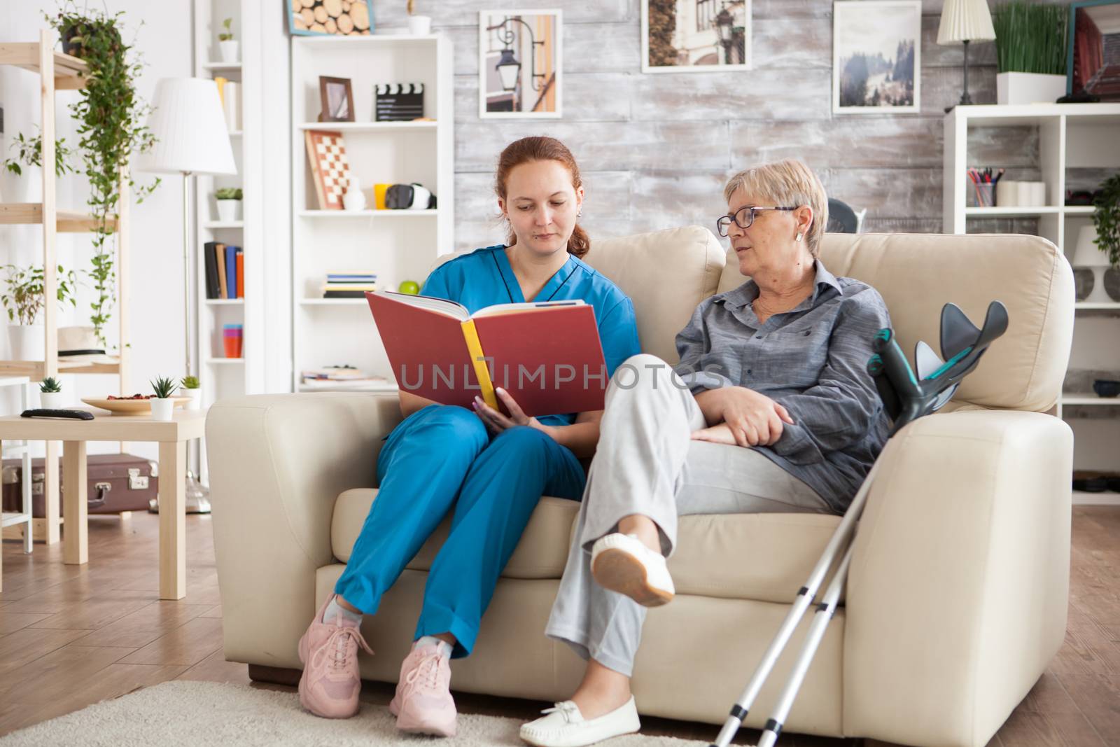 Young nurse sitting on couch in nursing home reading a book for old lady with crutches.