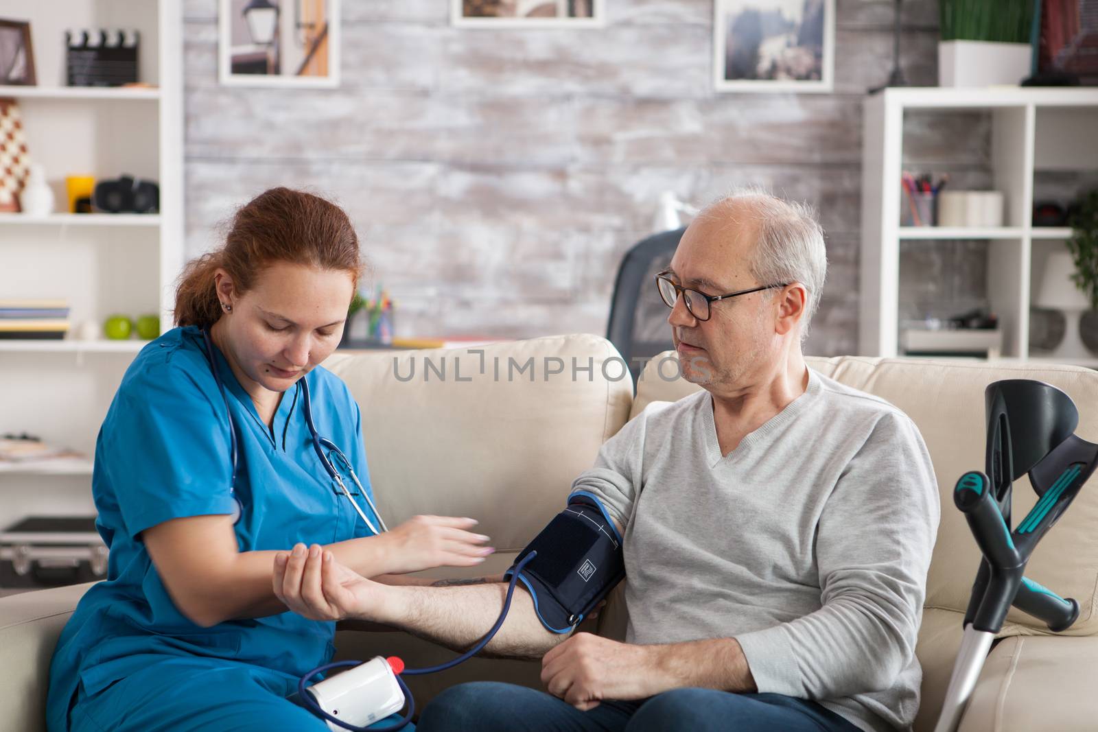 Retired old man sitting on couch in nursing home while female doctor is taking blood pressure using a digital device.