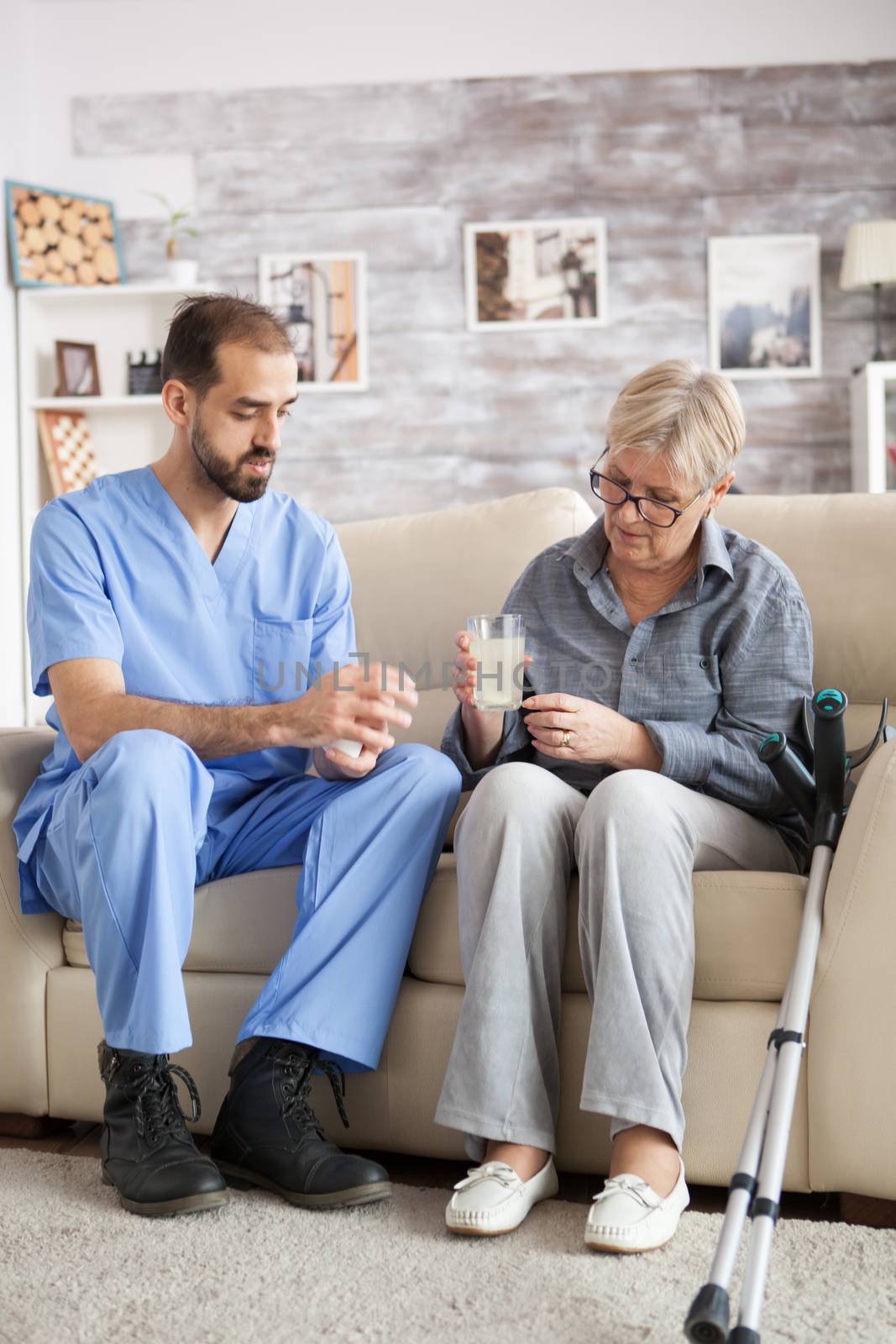 Male nurse sitting on couch with senior woman in nursing home helping her to take the medicine.