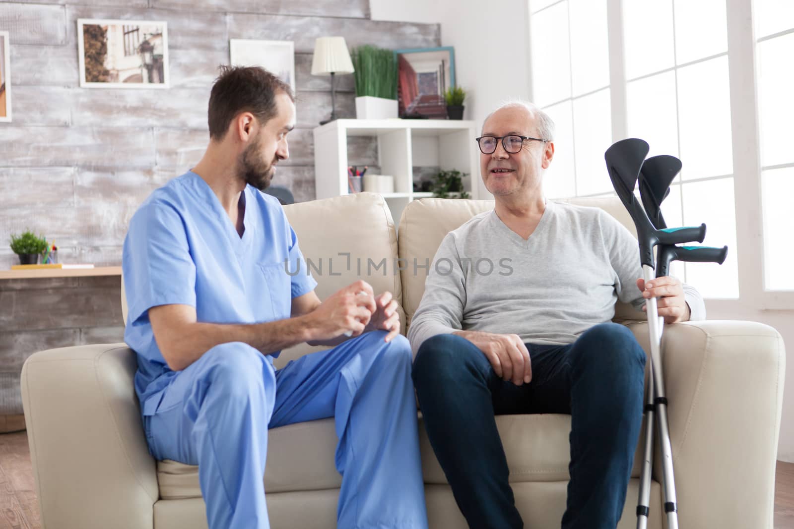 Elderly age man smiling while talking with male doctor in nursing home while holding crutches.