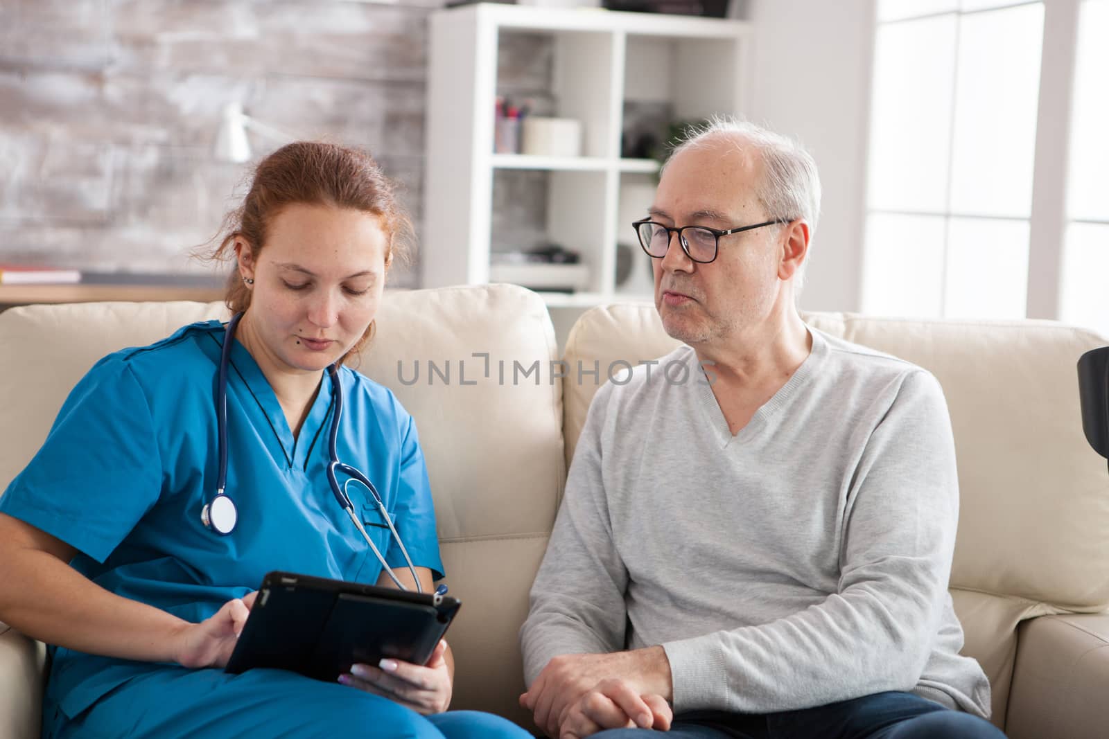 Young female doctor in nursing home taking notes on tablet pc while talking with senior man on the couch.