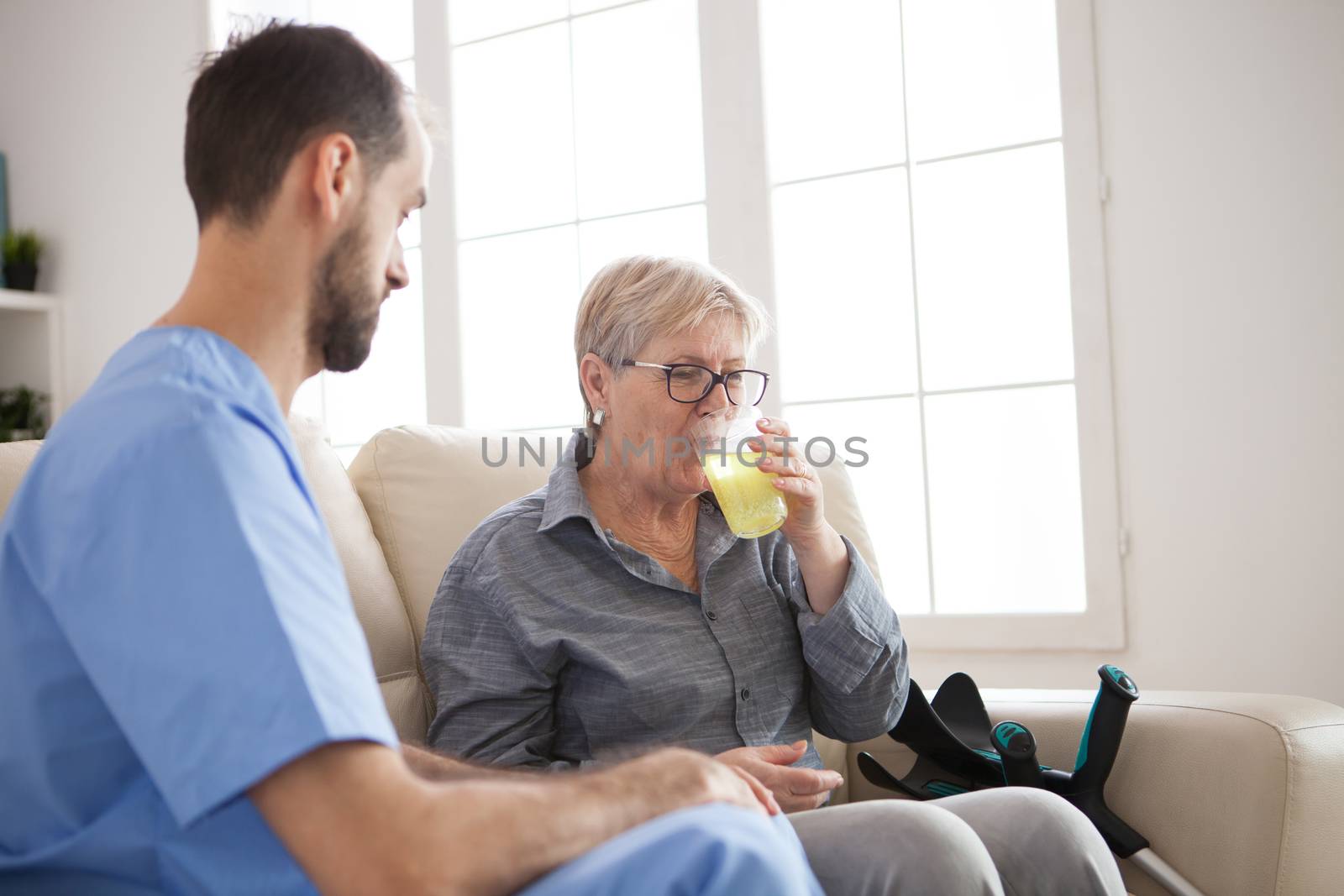 Young caregiver sitting down on couch with senior woman by DCStudio