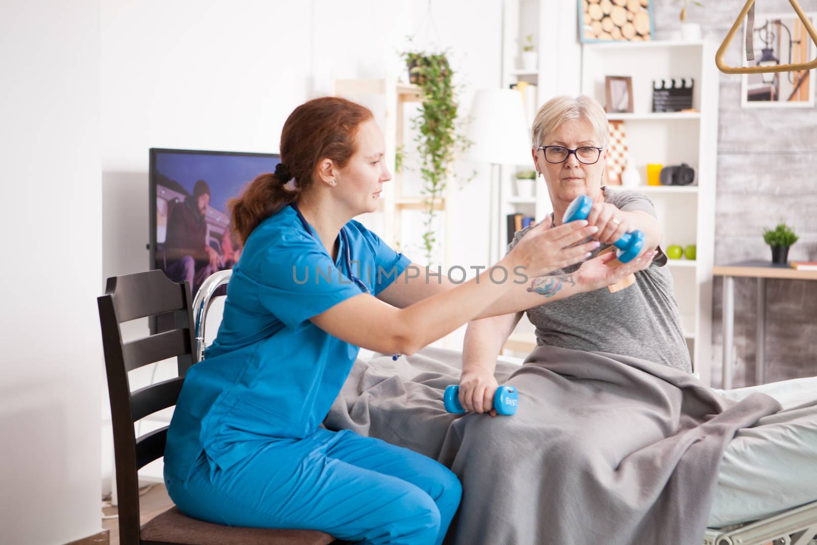 Senior woman in nursing home doing medical rehab using dumbbells with help from nurse.