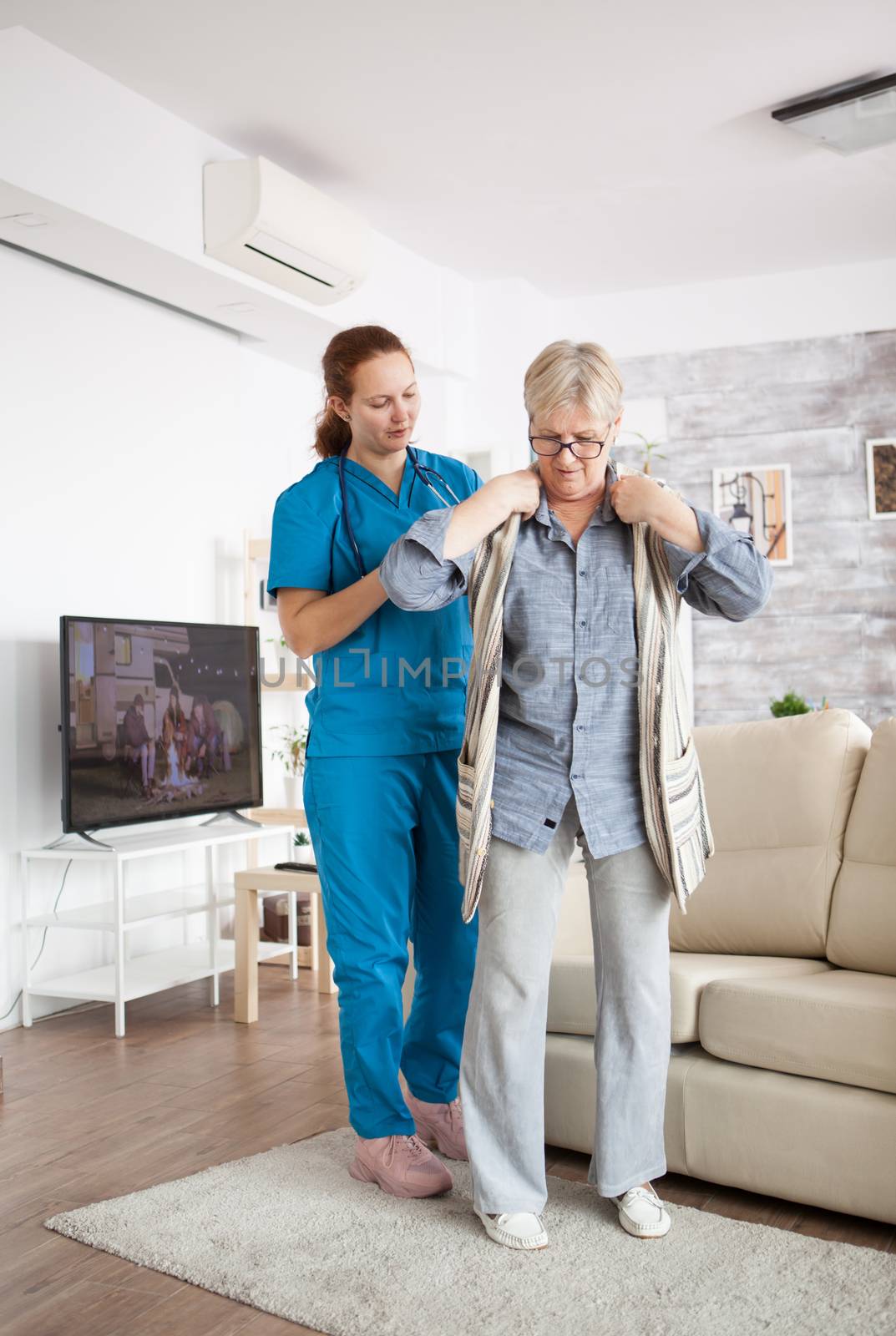 Young female nurse helping senior woman to get dressed in nursing home.